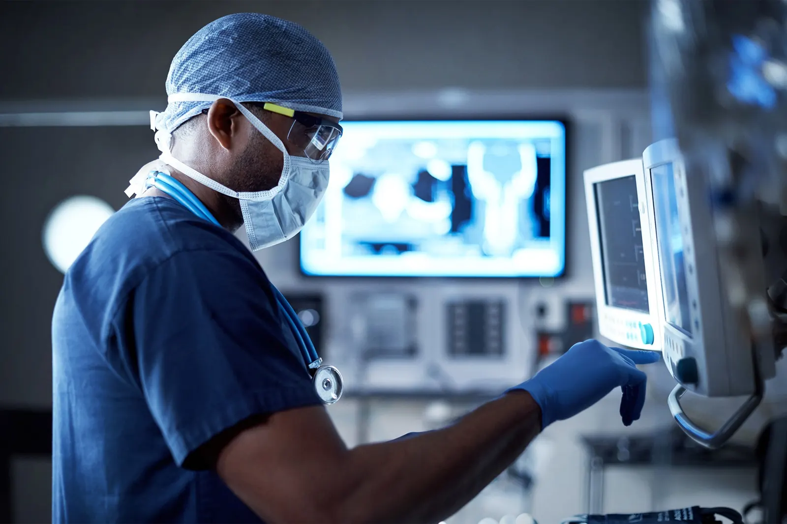 A clinician in surgical scrubs and a mask checks a patient monitor beside a wall of medical imaging